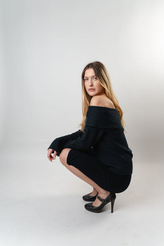 Model wearing the Amara Off-Shoulder Sweater in black knit, posing in a crouched position in a studio
