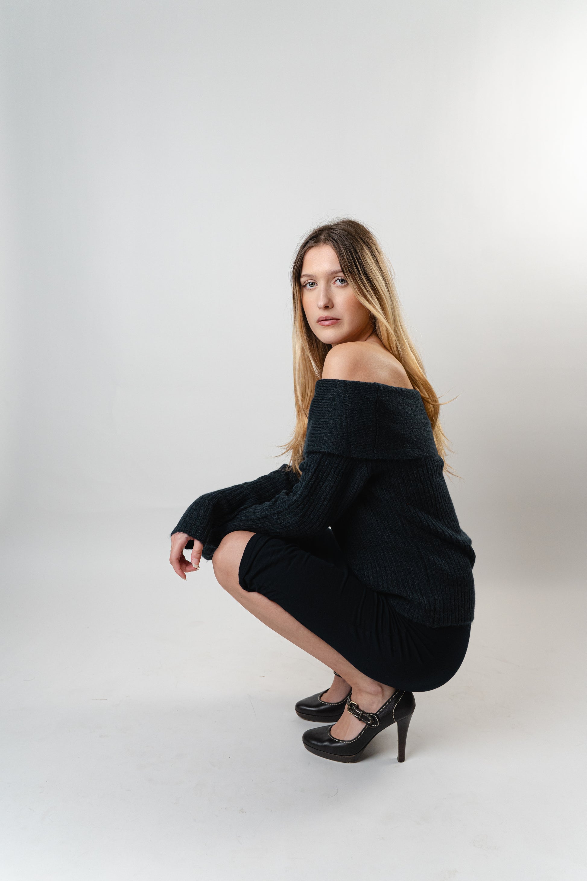 Model wearing the Amara Off-Shoulder Sweater in black knit, posing in a crouched position in a studio