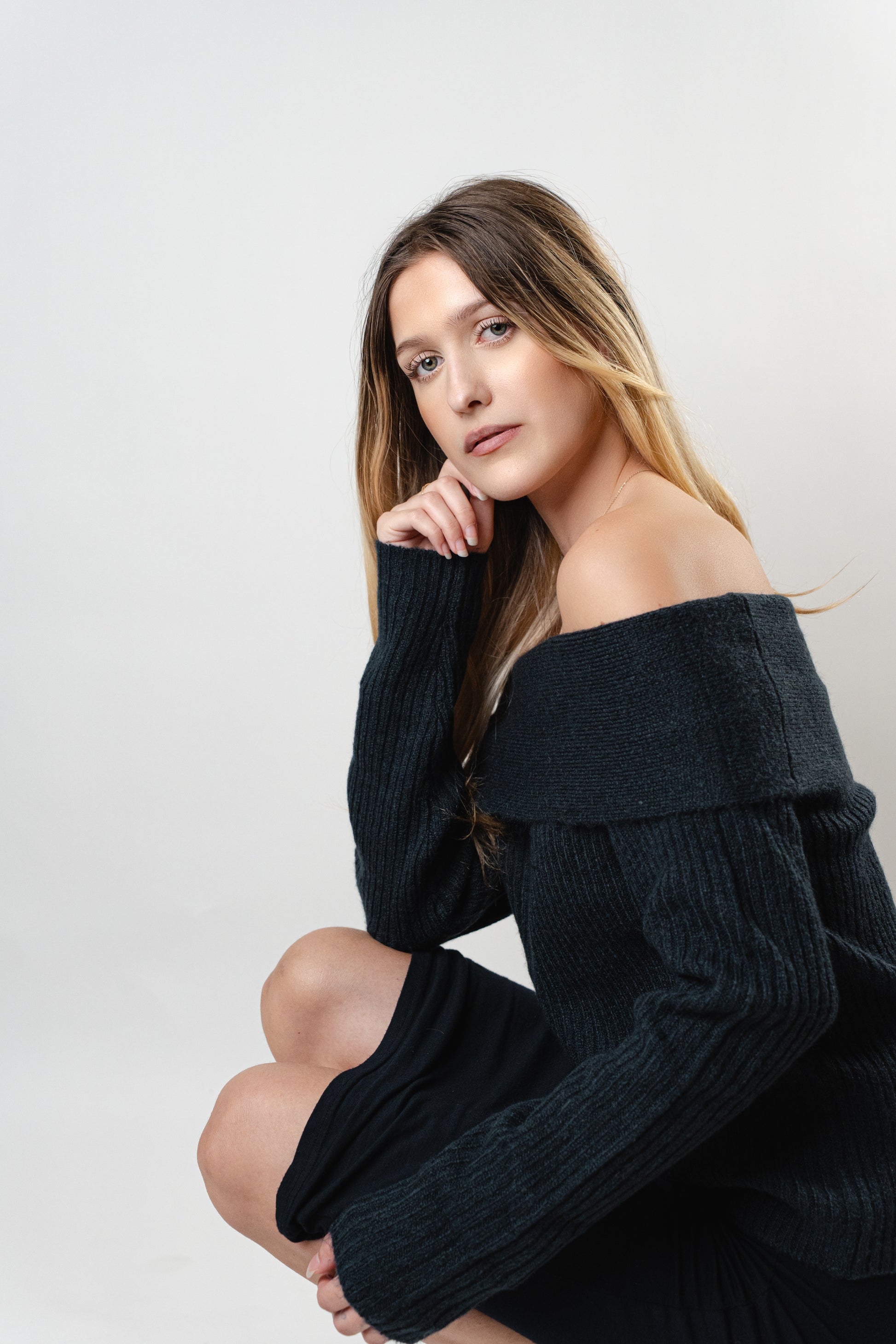 Model wearing the black Amara Off-Shoulder Sweater, posing seated in a studio with soft lighting.