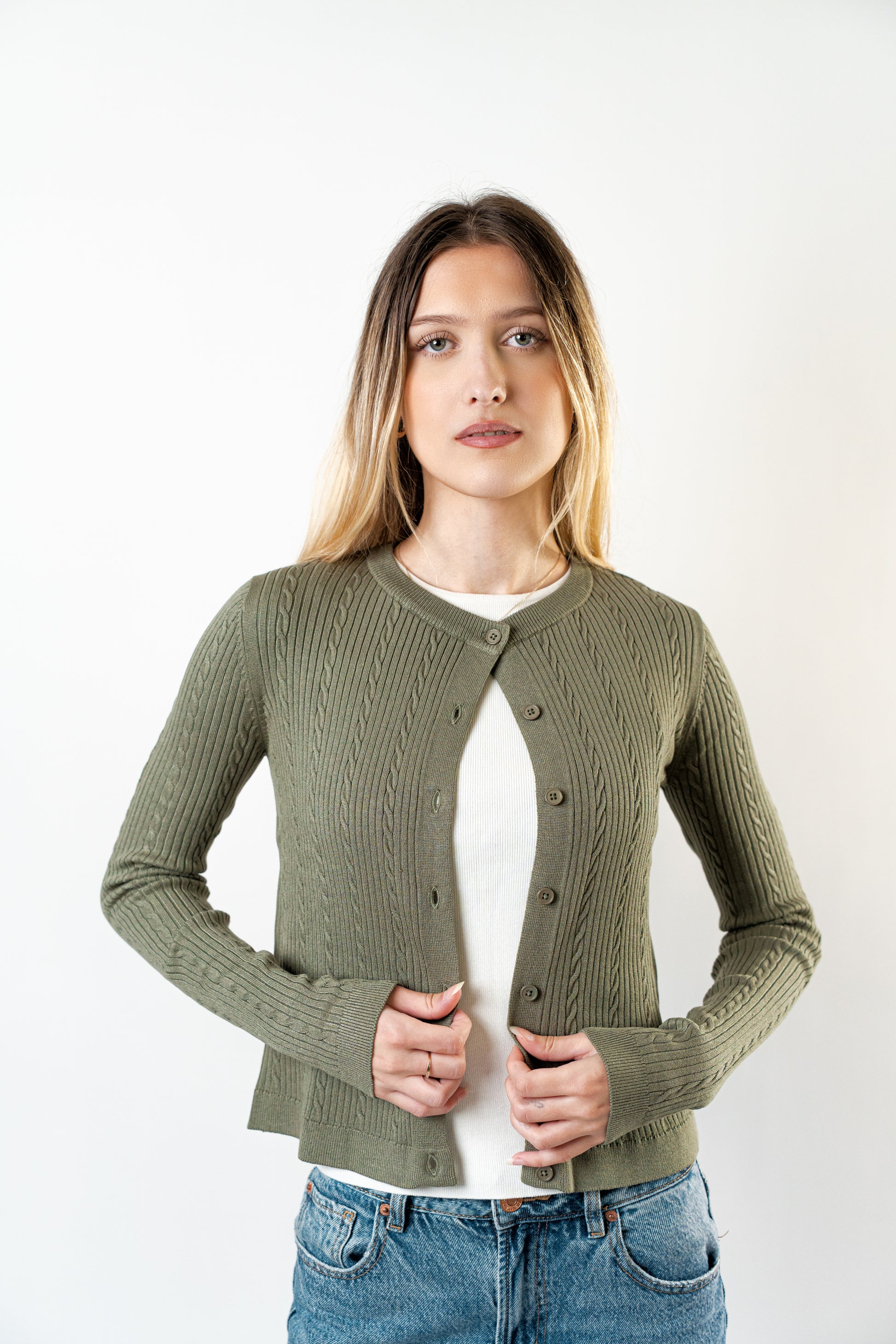 "Model adjusting the olive Celine Cable Knit Cardigan worn over a white ribbed top, showing textured knit details.