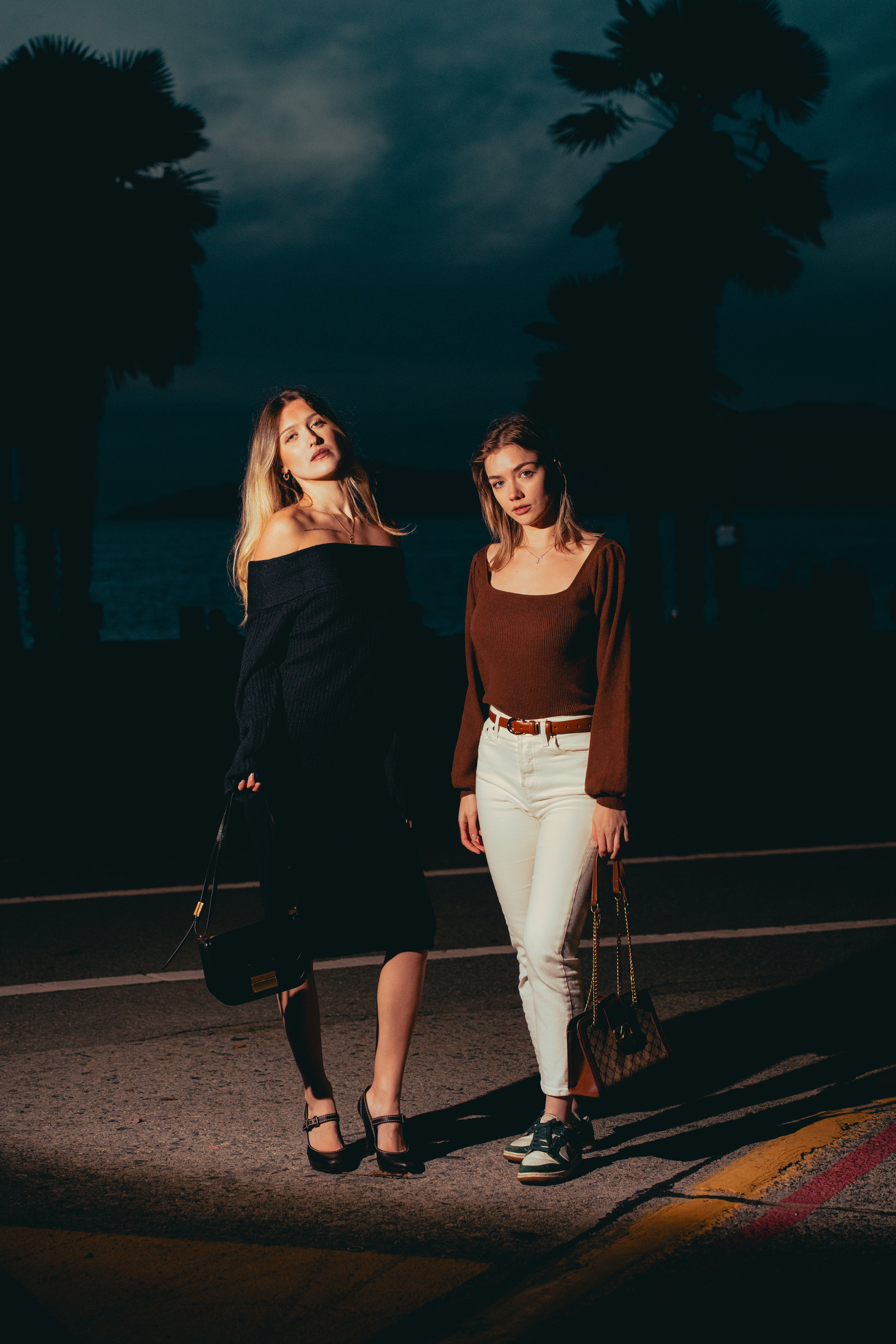 Two women standing on a street at night with palm trees in the background.